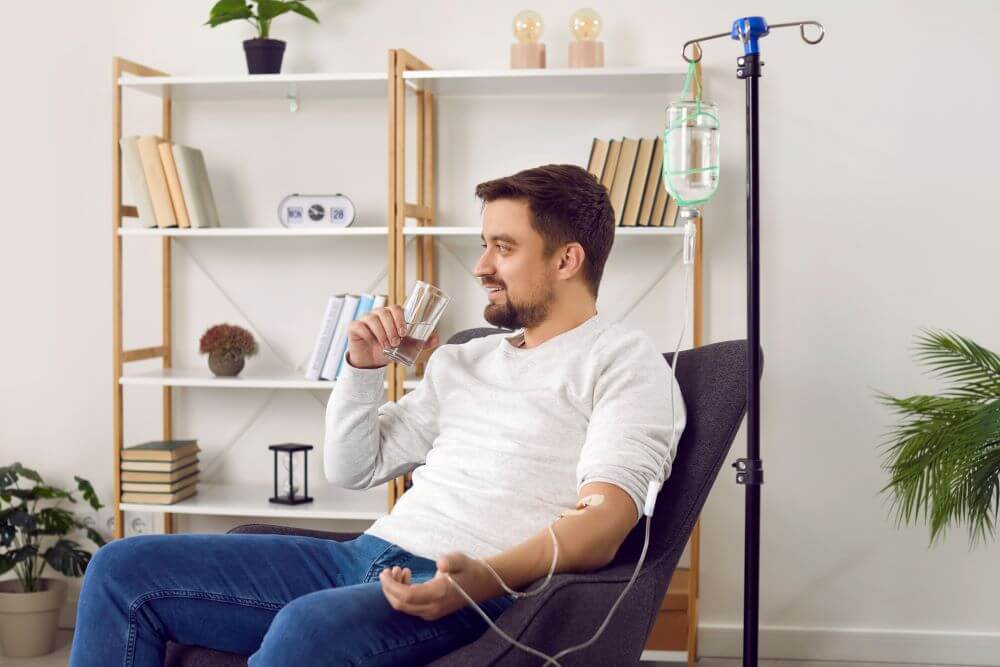 Bearded young man sitting in a chair drinking while having an IV drip to restore his hydration levels.